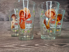 Vintage Coca Cola Magnificent Ladies Collector Series 1 Glasses featuring vibrant images of women in classic Coca-Cola advertising, displayed in a set of four on a wooden surface.