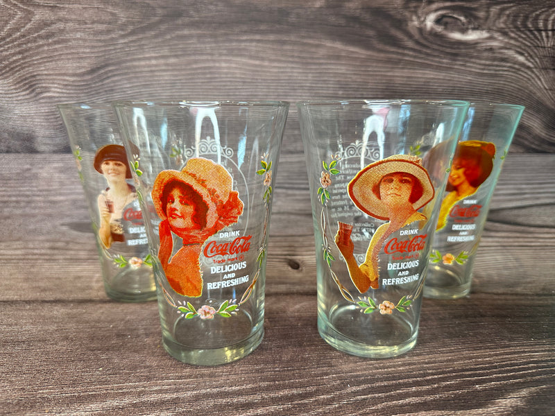 Vintage Coca Cola Magnificent Ladies Collector Series 1 Glasses featuring vibrant images of women in classic Coca-Cola advertising, displayed in a set of four on a wooden surface.