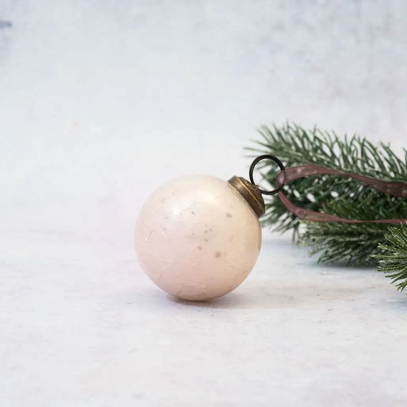 2" Alba Crackle Glass Christmas Bauble in a soft pink hue, featuring a bronze cap and brown organza ribbon, displayed next to a frosted pine branch on a neutral background.