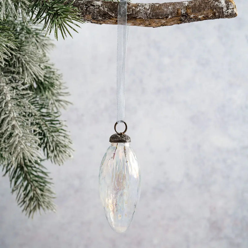 2" Clear Luster Conical Glass Christmas Ornament with an iridescent finish, bronze cap, and white organza ribbon, hanging from a branch with a pine tree in the background.