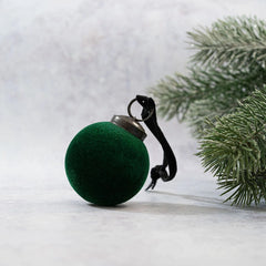 2-inch emerald velvet-covered glass hanging bauble with an ornate bronze cap and matching ribbon, resting beside evergreen branches for a festive holiday display.