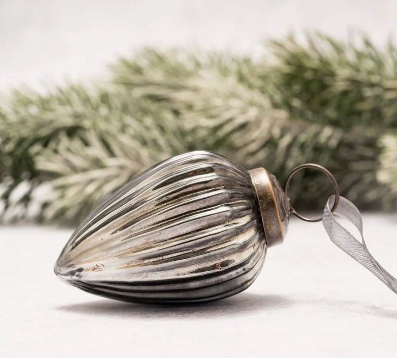 2" Medium Slate Ribbed Glass Pinecone Ornament featuring a ribbed texture, ornate bronze cap, and coordinating organza ribbon, lying beside frosted greenery.