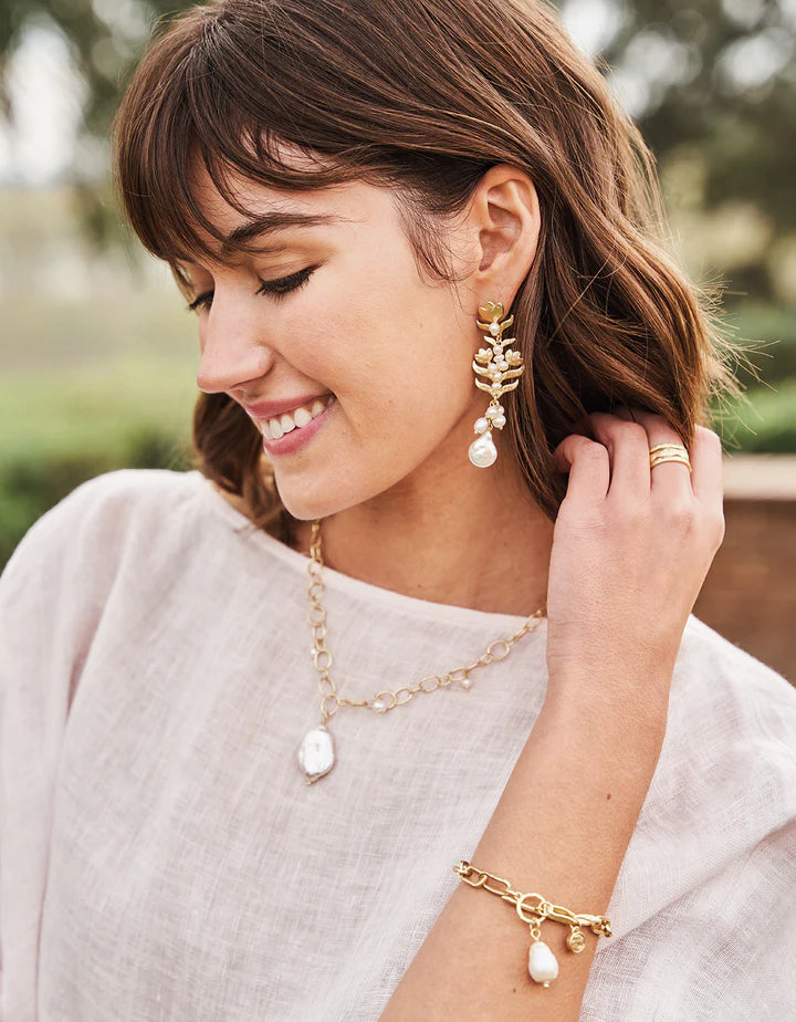 Woman smiling and wearing matching gold jewelry, including a freshwater pearl necklace, pearl drop earrings, and a gold toggle bracelet with a freshwater pearl charm, styled with a light blouse.