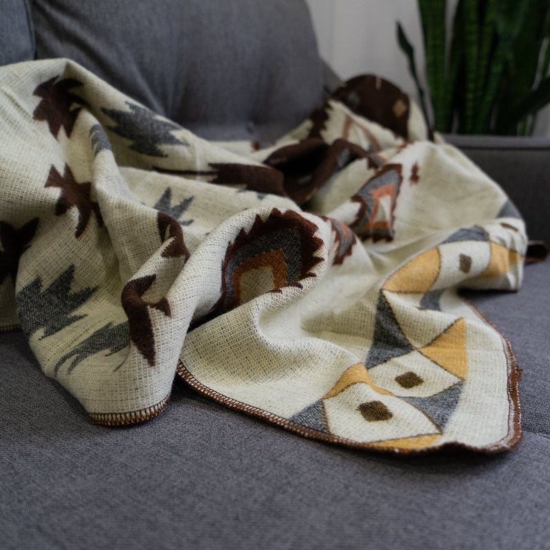 Andean Alpaca Wool Throw Blanket in a cream colorway, draped over a grey sofa, showing a pattern of stylized brown and gray leaf/feather shapes and yellow-accented geometric owl motifs.