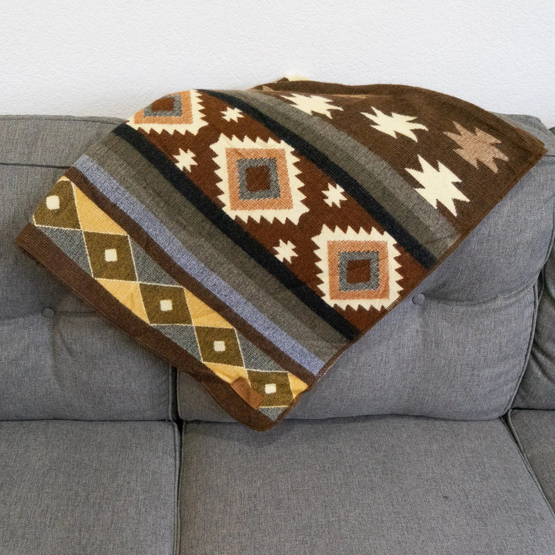 Close-up of a folded Andean Alpaca Wool Throw Blanket on a gray sofa, highlighting the striped tribal pattern with white diamond shapes, brown star motifs, and bands of gold, gray, and blue.
