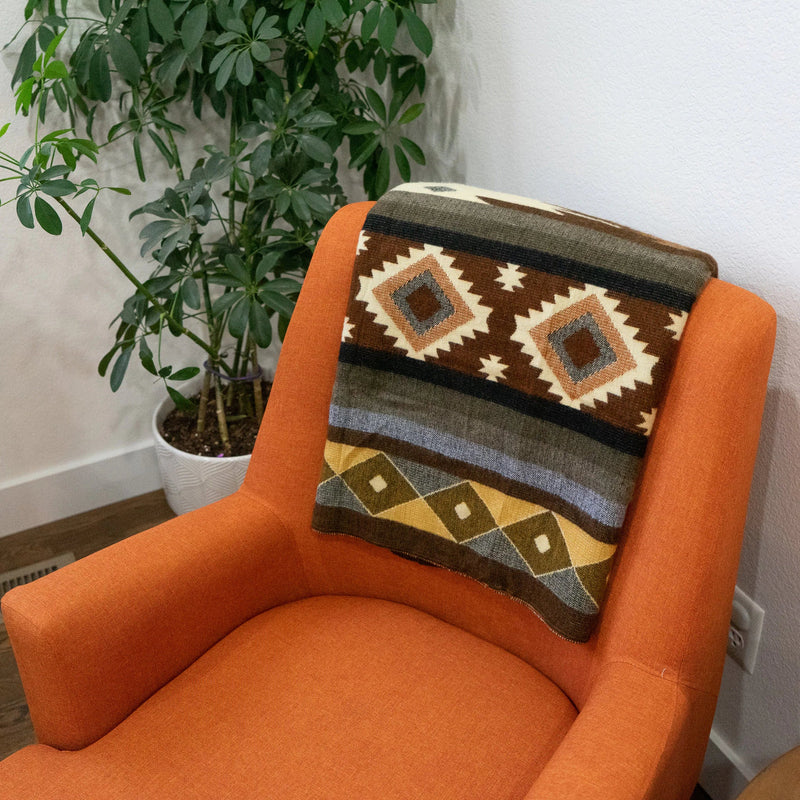 Andean Alpaca Wool Throw Blanket in the Mojave geometric pattern, draped over the back of a brightly colored orange mid-century accent chair next to a large potted plant.