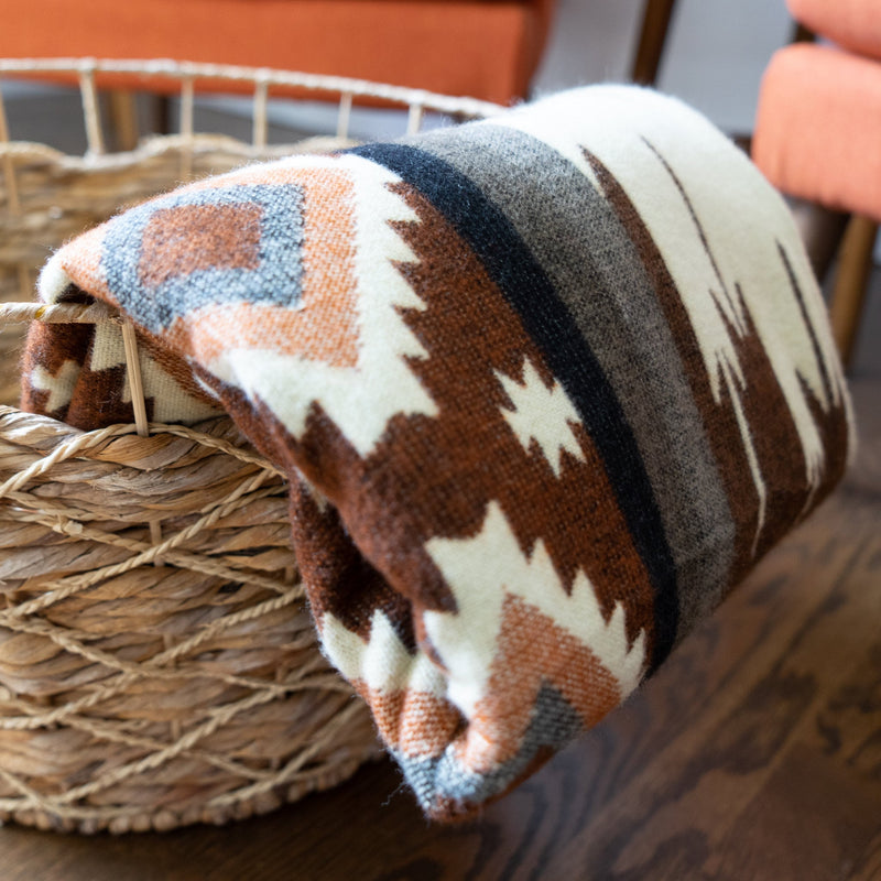 Andean Alpaca Wool Throw Blanket featuring a white and brown tribal stripe pattern, neatly folded and stored inside a natural woven wicker basket on a hardwood floor.