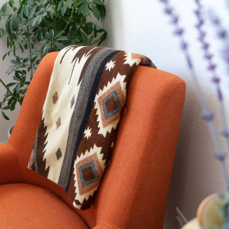 Andean Alpaca Wool Throw Blanket in a white, brown, and gray tribal geometric pattern, draped over the back of a bright orange accent chair next to a potted plant.