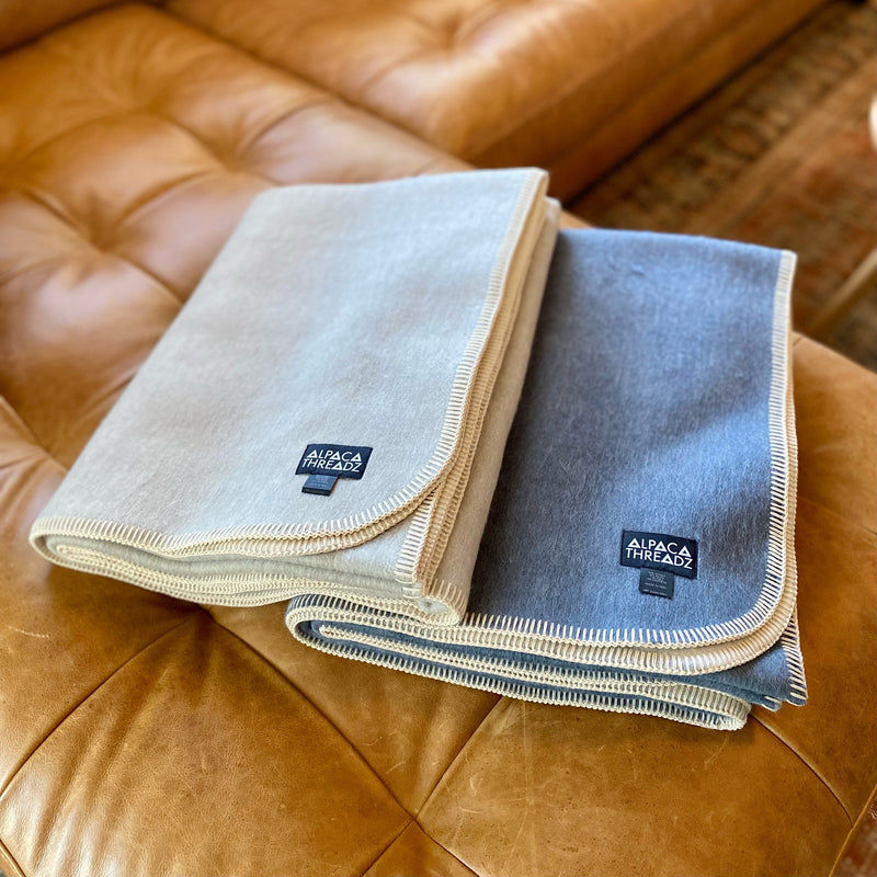 Two folded Alpaca Camp Throws, one in beige/light tan and one in charcoal gray, featuring blanket stitch edging and a woven label, resting on a brown leather couch.