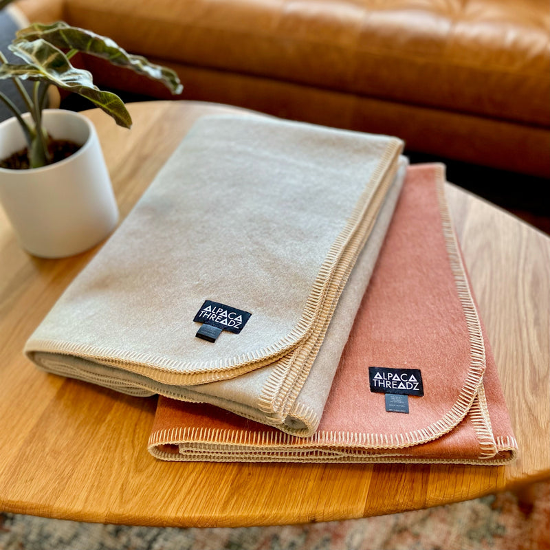 Two folded Alpaca Camp Throws in beige/tan and burnt rose/terracotta, featuring 'Alpaca Threadz' label and blanket stitch edging, stacked on a wooden side table in a living room