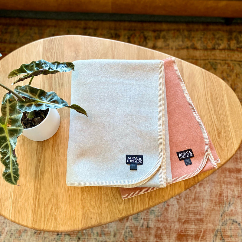 Two folded Alpaca Camp Throw blankets in light grey and salmon shades displayed on a wooden table beside a small potted plant, showing their soft texture and stitched edges.