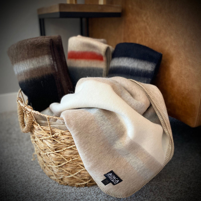 Assorted alpaca camp throws in tan, red, brown, and grey tones displayed in woven basket – soft premium coastal home décor accent