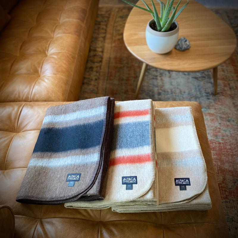 Stack of three alpaca camp throws in brown, red, and cream tones on leather sofa – premium coastal home décor accent
