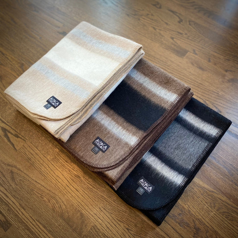 Set of three alpaca camp throws in cream, brown, and grey tones folded on wooden surface – premium coastal home décor accent