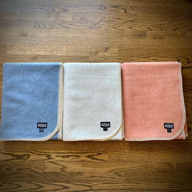 Three folded Alpaca Camp Throw blankets in sky blue, light grey, and salmon shades neatly arranged on a wooden surface, showcasing their smooth alpaca texture and stitched edging.