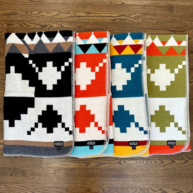 Four geometric alpaca wool blankets stacked side-by-side on wood, showing Black/White, Red/White, Blue/Yellow, and Green/White colorways.