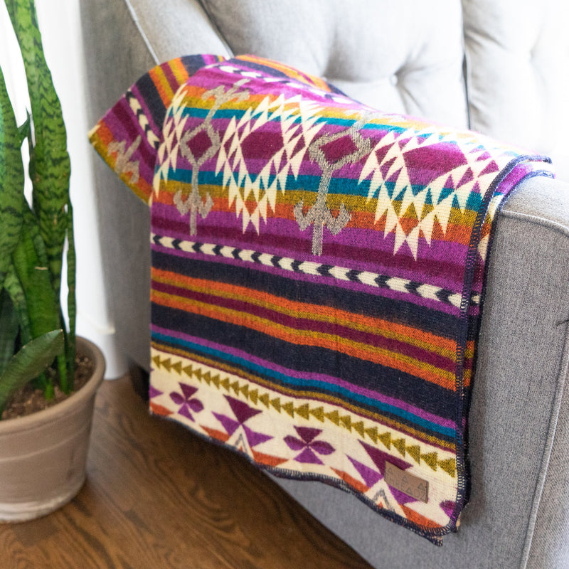 Corner view of the Amethyst alpaca blanket draped on a sofa in a bright interior, showing the vivid jewel tones.