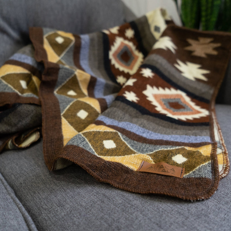 Andean Alpaca Wool Throw Blanket in Mojave Pattern, folded on a grey sofa, showing the rich brown, gold, cream, and blue geometric design and the leather brand tag.