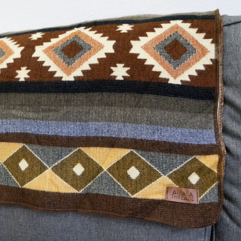 Close-up detail of the Andean Alpaca Wool Throw, showing horizontal stripes of brown, gray, and blue, with large diamond patterns and the "Alpaca Threadz" leather brand tag.