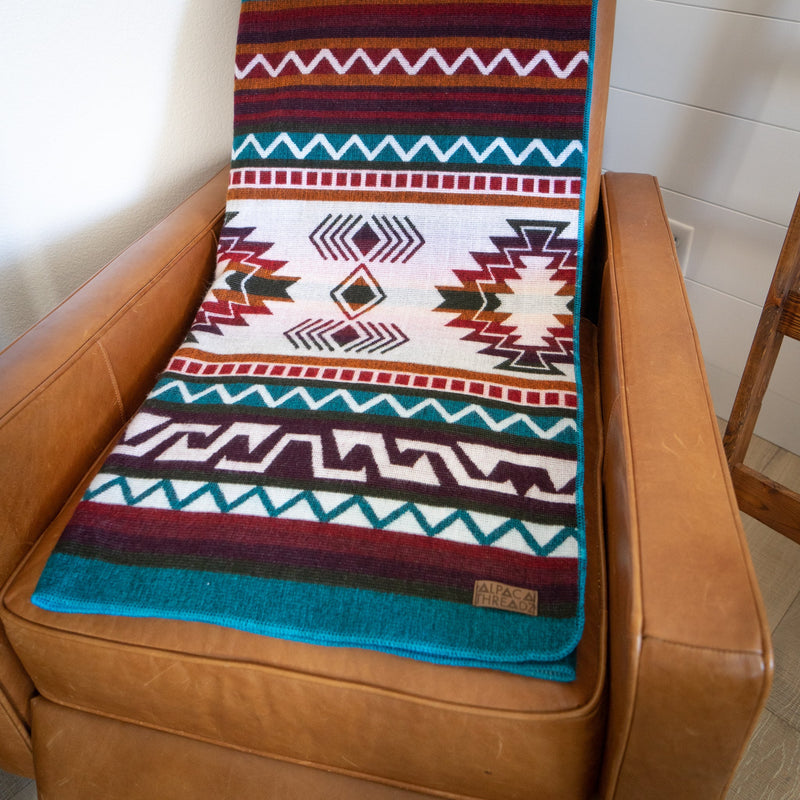 Turquoise Andean alpaca wool blanket with colorful geometric design draped over tan leather chair – vibrant artisanal coastal home décor accent