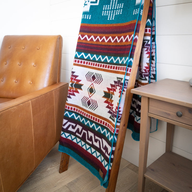 Turquoise Andean alpaca wool blanket with colorful geometric pattern draped on wooden ladder beside leather chair – artisanal coastal home décor accent
