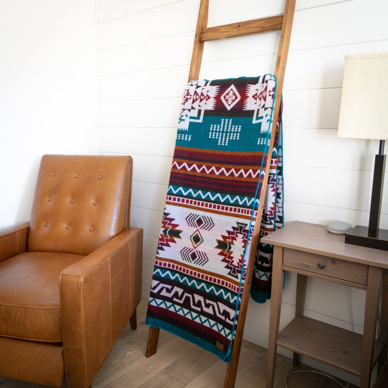 Turquoise Andean alpaca wool blanket with colorful geometric pattern draped on wooden ladder beside tan leather chair – vibrant coastal home décor accent