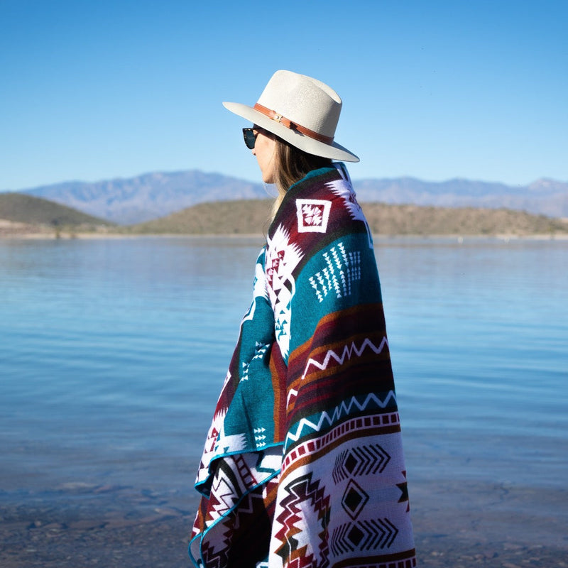 Person wearing turquoise Andean alpaca wool blanket with colorful geometric pattern standing by the water – artisanal coastal lifestyle décor accent