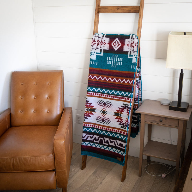 Turquoise Andean alpaca wool blanket with vibrant geometric design draped on wooden ladder beside leather chair and lamp – artisanal coastal home décor accent