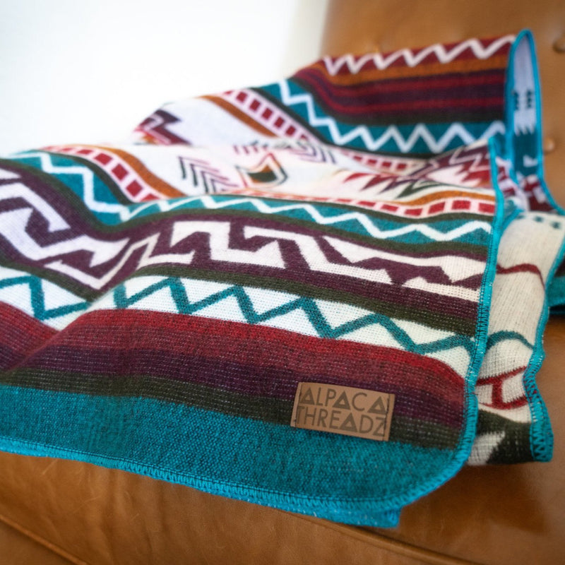 Close-up of turquoise Andean alpaca wool blanket with colorful geometric tribal pattern draped on tan leather chair – soft artisan-made textile décor