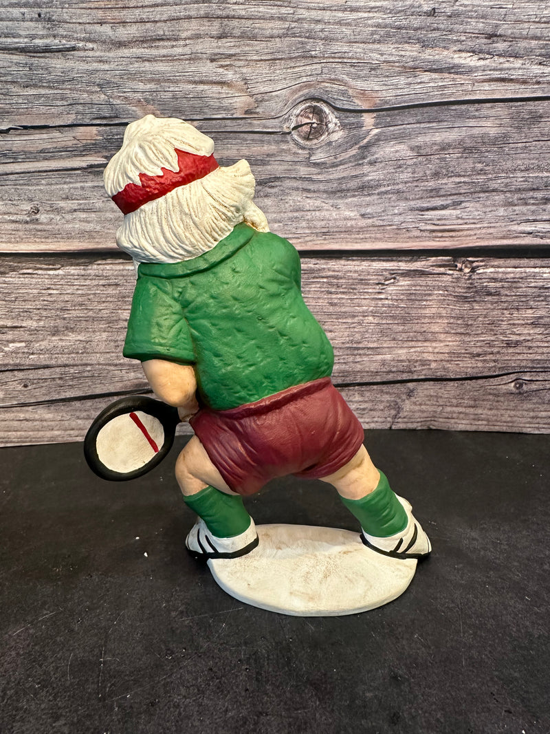 Santa figurine playing pickleball, wearing a green shirt, red shorts, and a red headband, holding a paddle with a yellow pickleball, standing on a white base with a wooden background.