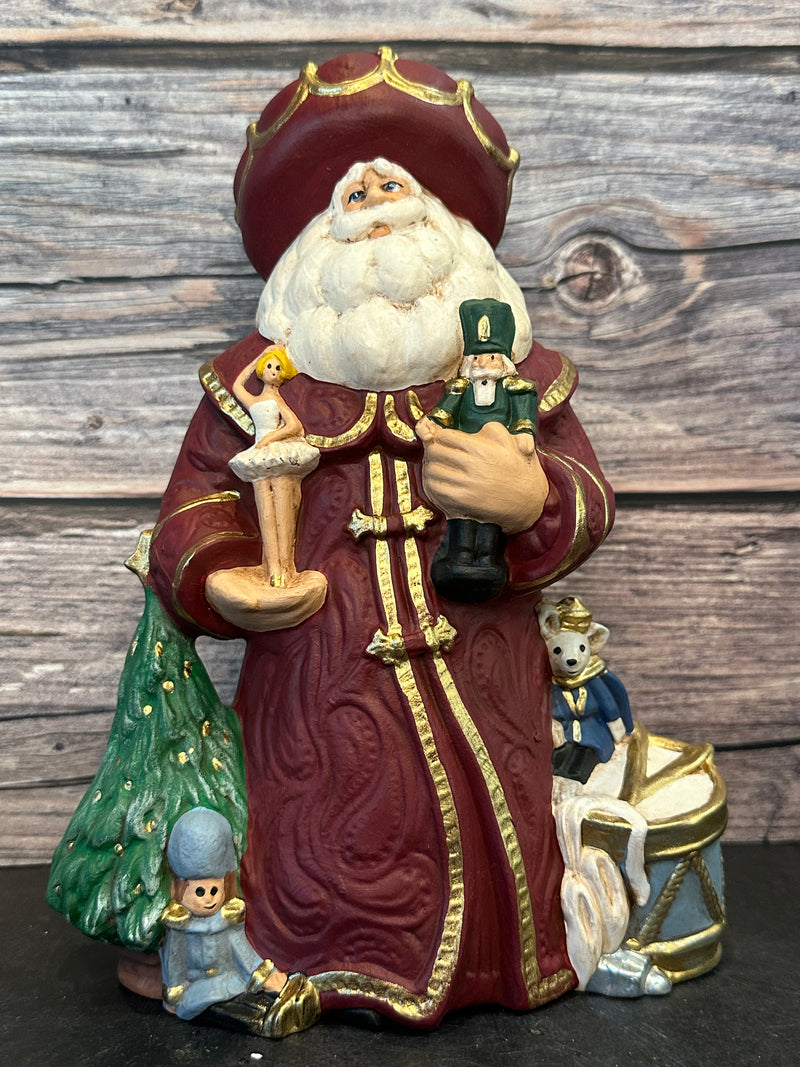 Ceramic Santa figurine in a burgundy robe, holding a nutcracker and ballerina, surrounded by ballet slippers, toy soldiers, and a Christmas tree, with intricate gold accents.