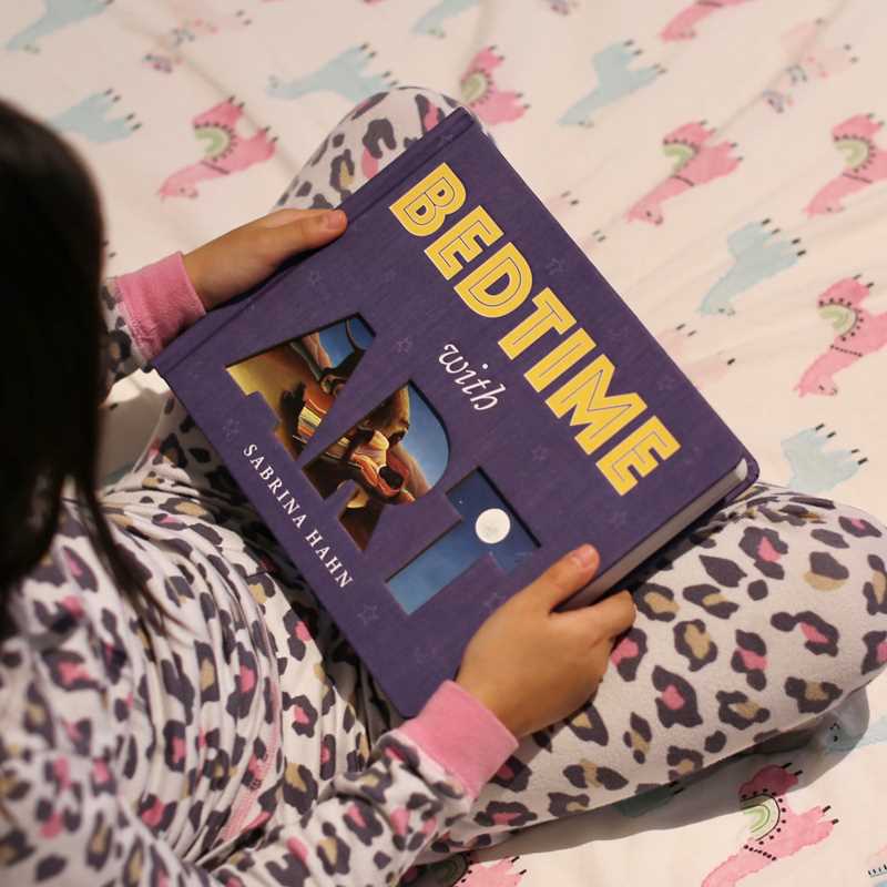 Person holding a book titled 'Bedtime with Sabrina Hahn' on a patterned bedspread.