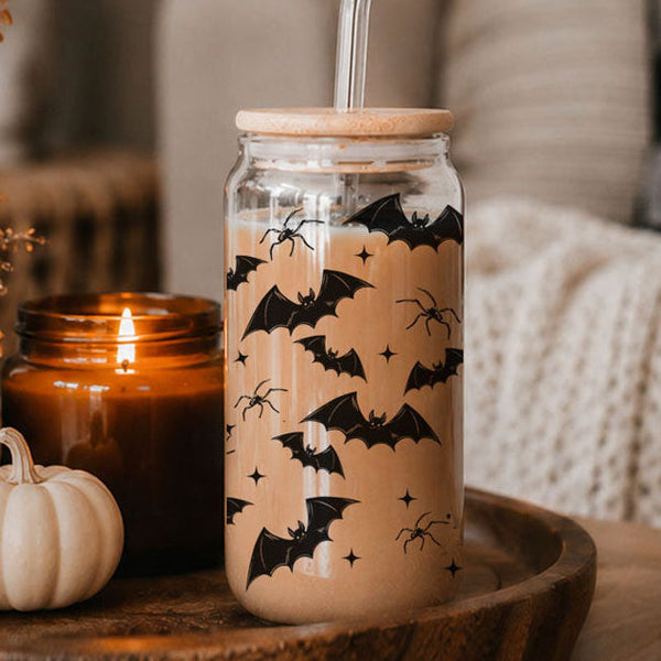 Clear glass tumbler with black bat graphics, bamboo lid and glass straw beside an amber candle and white pumpkin.