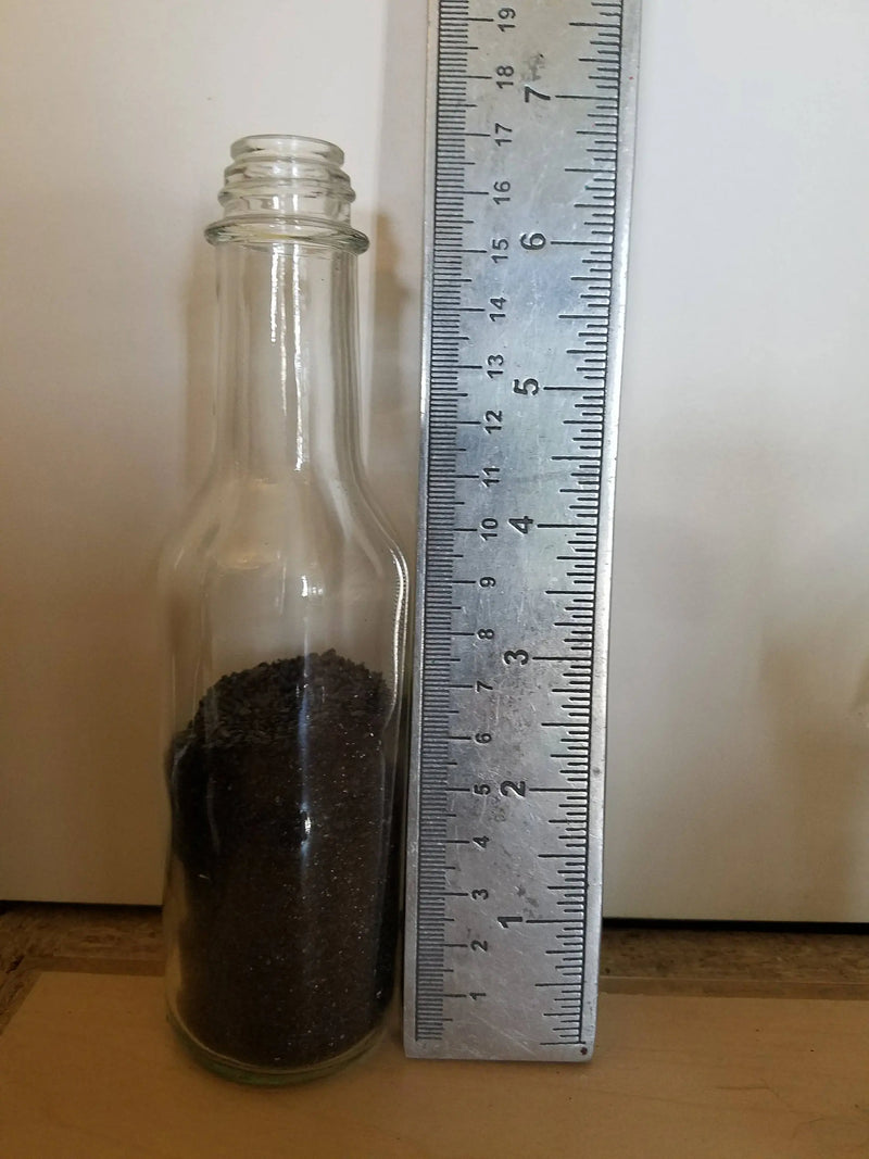 Clear glass bottle filled with Black sand next to a ruler on a white background