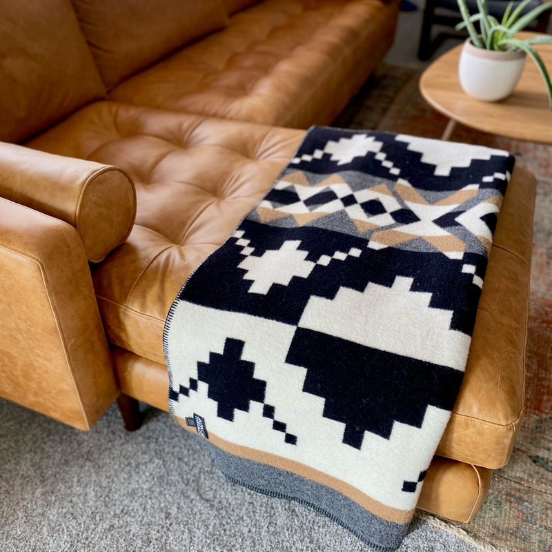 Black and white geometric alpaca throw blanket draped casually over a brown leather sofa in a cozy interior setting.