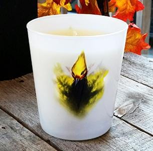 White frosted glass candle featuring a yellow and black bird design on the front, displayed on a wooden surface with autumn leaves in the background.