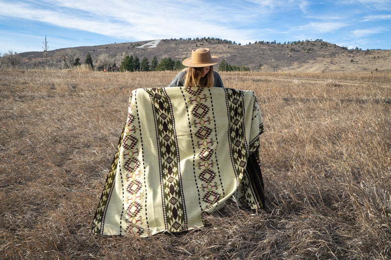 Woman sitting on the ground with the Cactus Alpaca Blanket draped around her, showing cozy outdoor use.