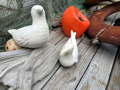 Two white ceramic seagull sculptures on a wooden deck with coastal fishing net and nautical décor in the background.