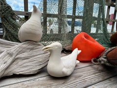 Close-up of two white ceramic seagulls placed on a weathered driftwood piece with a vibrant orange buoy in the background.