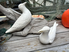 White ceramic seagulls sitting on driftwood, surrounded by nautical fishing net and wooden decor, showcasing their detailed feather patterns.
