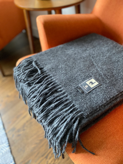Charcoal gray Tumi Alpaca Wool Throw Blanket folded neatly on an orange chair, featuring soft fringe detailing and a stitched brand label for a refined, cozy finish.