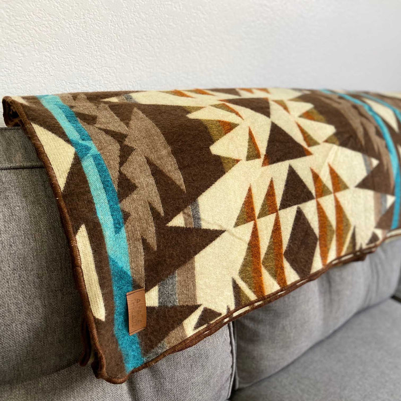 The Cowboy Andean Alpaca Wool Blanket draped on a grey couch, showcasing the rich pattern featuring brown, charcoal, white, and turquoise accents.