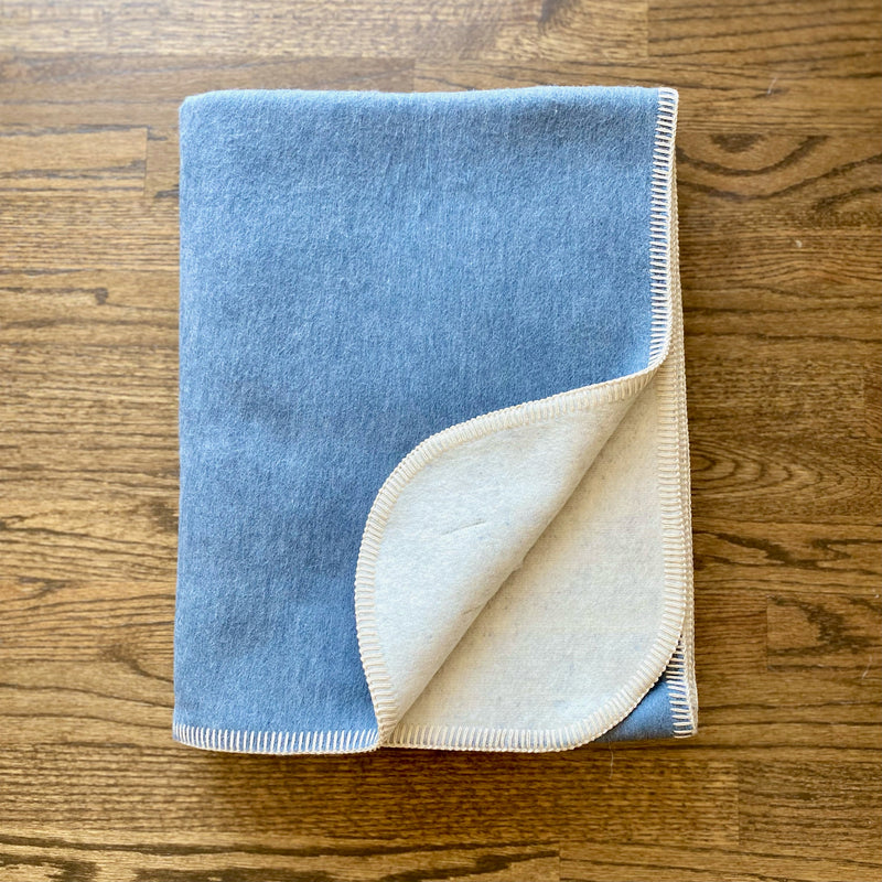 Reversible blue fleece and off-white sherpa throw blanket with visible cream blanket stitch edging, folded on a wooden floor