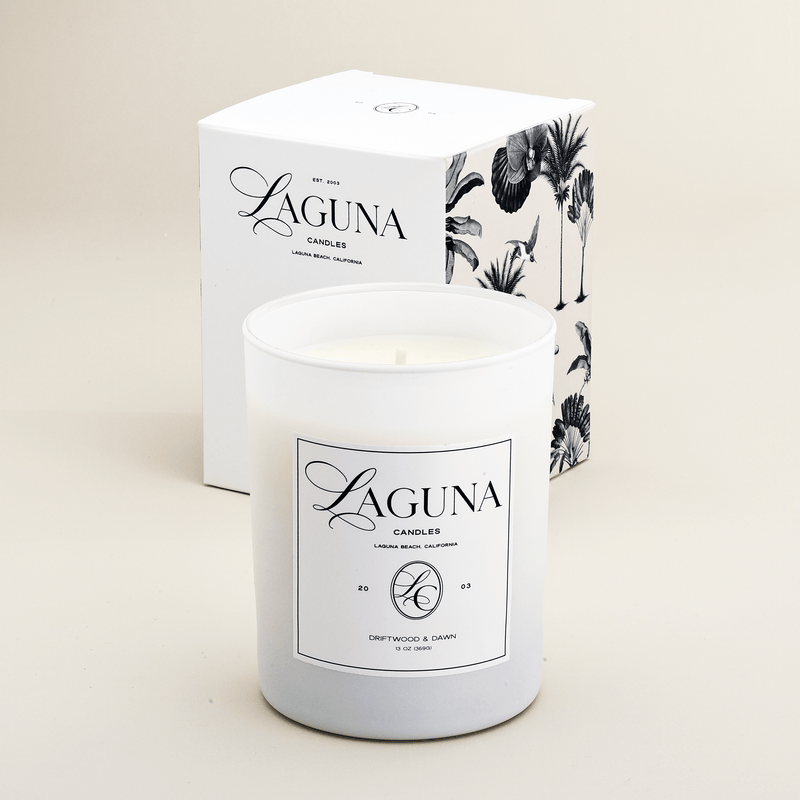 Studio shot of the Laguna Candles Driftwood & Dawn 13oz candle in a white frosted vessel next to its white box with black tropical palm illustration.