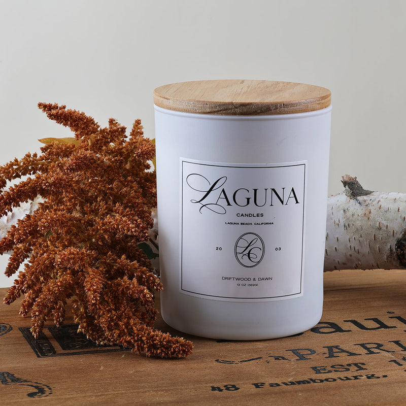 Styled shot of the Driftwood & Dawn 13oz candle with a natural wood lid, accented by warm reddish-brown feathery botanicals and a cozy background.