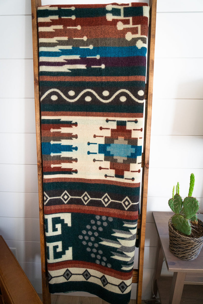 Evergreen Alpaca Blanket on a ladder beside a side table with a lamp and cactus, showing it as a vertical home accent piece.