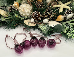 Five mulberry-colored crackle finish baubles laid out in a line with their organza ribbon ties, displayed against a decorative holiday wreath with seashells and pinecones.