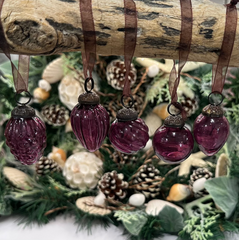 Mulberry crackle glass baubles hanging from brown ribbons on a rustic branch, with a festive backdrop of holiday greenery, pinecones, and seashells.