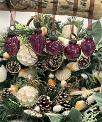 Close-up view of mulberry-colored crackle glass baubles hanging from a branch, surrounded by a lush holiday display of seashells and pinecones.