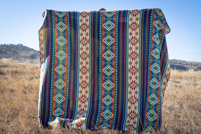 Galapagos Alpaca Blanket held up and spread wide, showcasing the full pattern of colorful vertical stripes in an open, grassy field.
